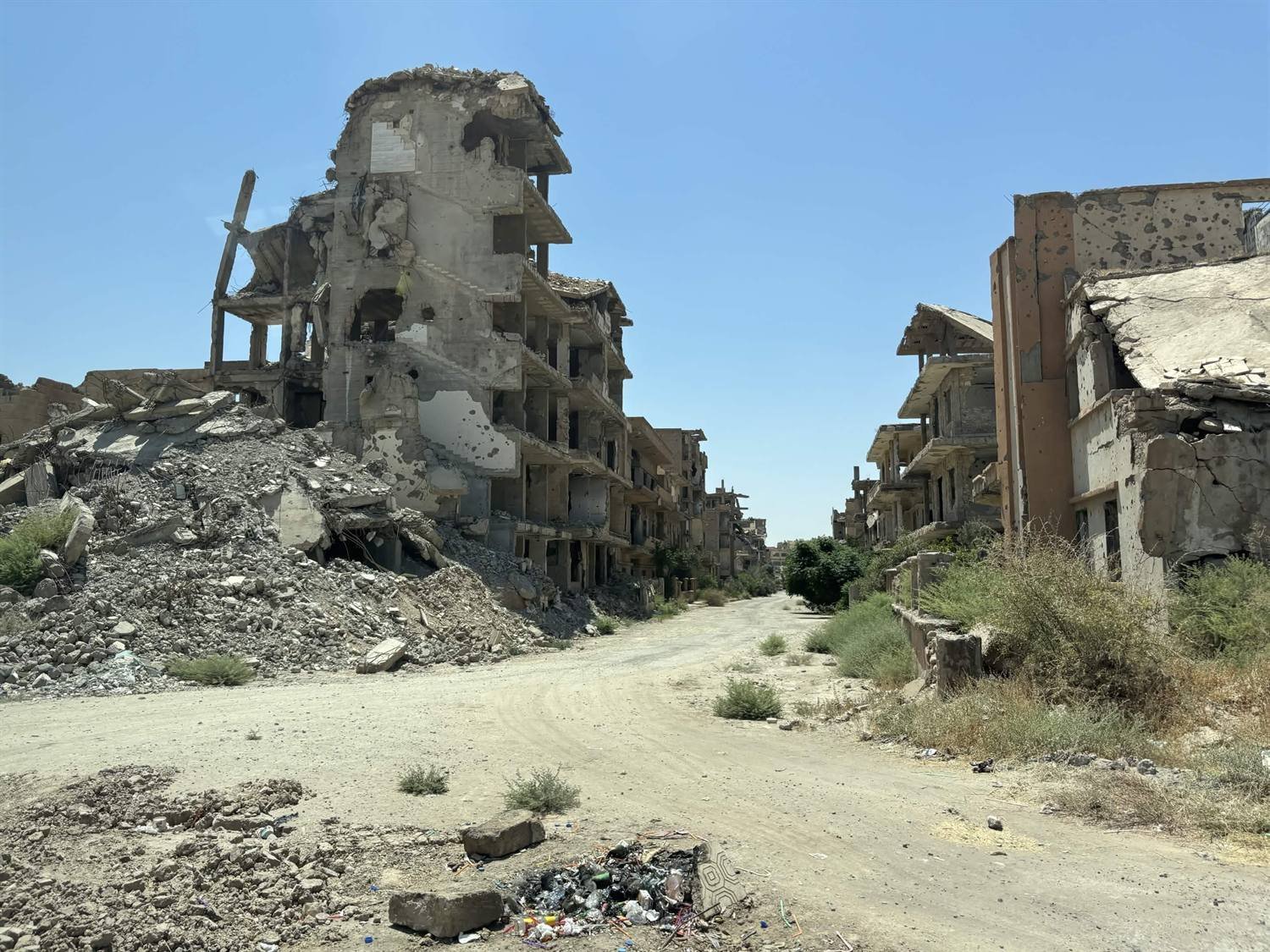 A street in Deir ez-Zor lined with destroyed and shell-damaged apartment blocks, with piles of rubble and a dirt track running between them.