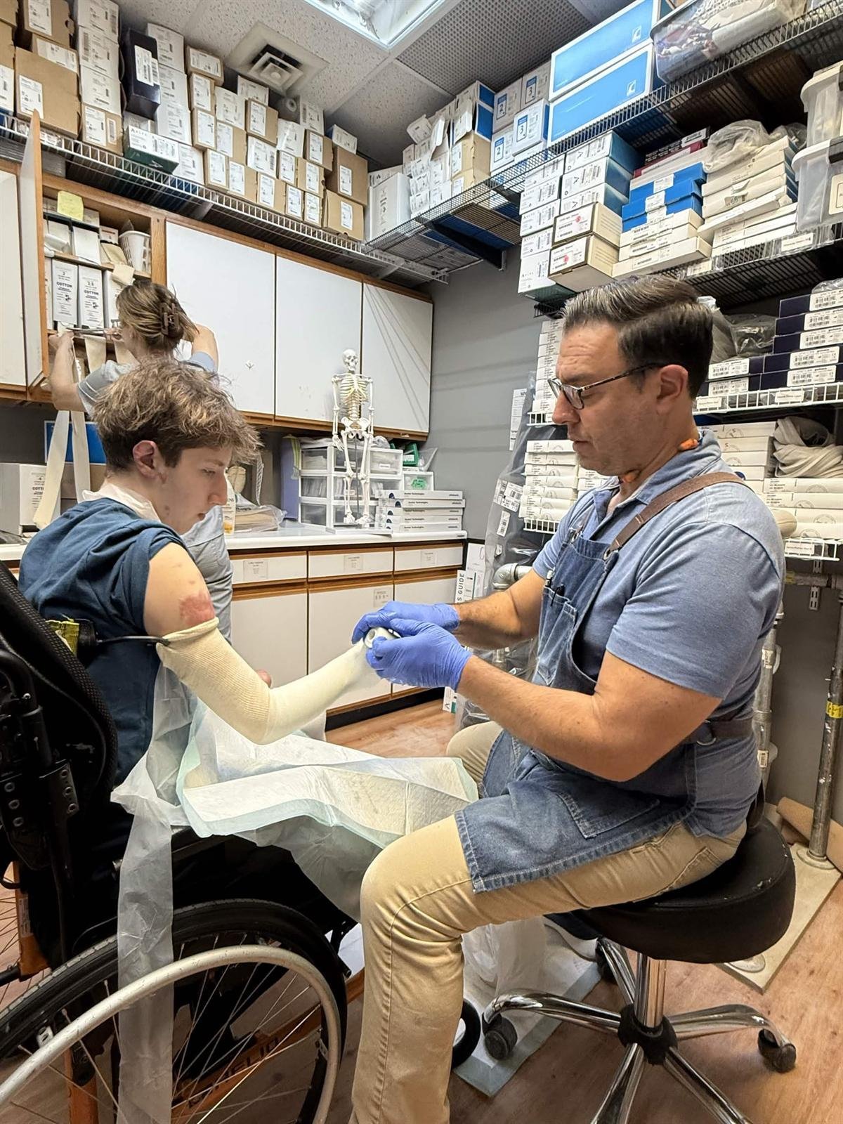 Co-founder Sam Lababidy seated in a wheelchair while a prosthetist fits a cast to his residual limb in a clinic lined with supplies.